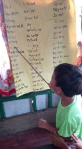 The image shows Charlie using a stick to point to words written on Manila paper. He is teaching his classmates how to read.